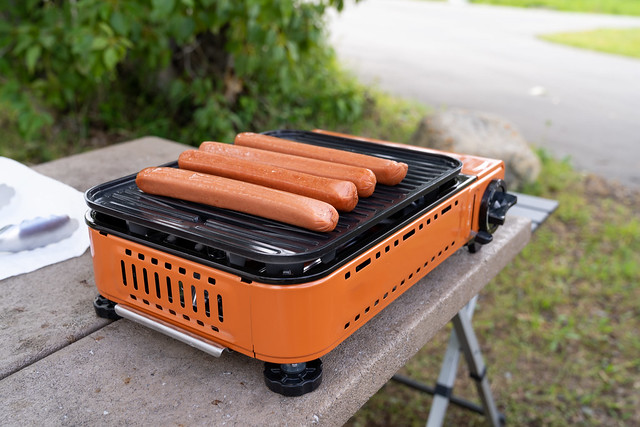 Hot dogs cook on an outdoor camp stove grill at a picnic