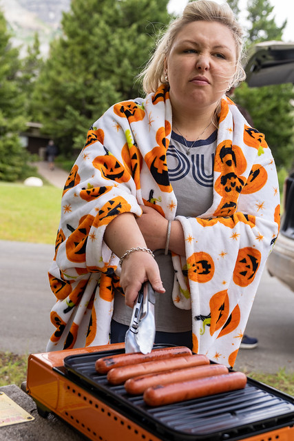 Angry, irritated blonde woman cooks and grills hot dogs at a picnic ground outside, wearing a pumpkin blanket