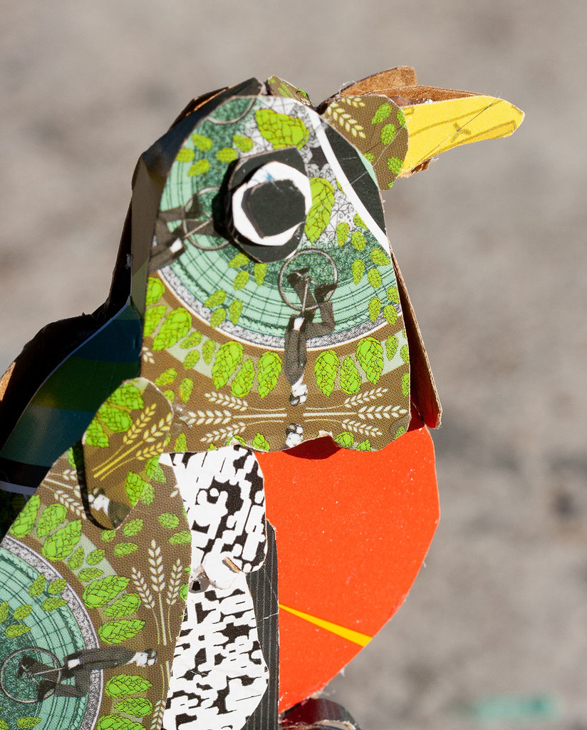Detail view of the head of the sculpture, the beak is yellow, protruding from an extra piece of green cardboard, also with men on unicycles woven into the green pattern of the beer box