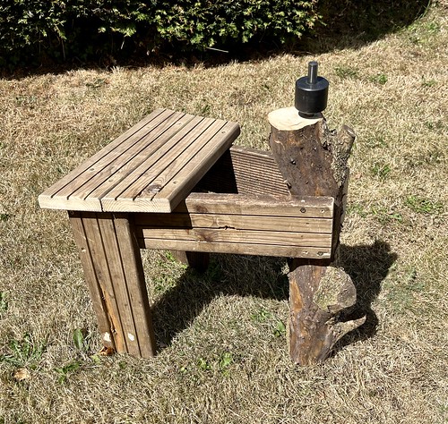 A photo of a small stool-like thing made out of wooden decking. At the front is a small log, positioned vertically as the third leg of the stool, with the top higher than the stool's seat. In the top of the log is a small metal cylinder.