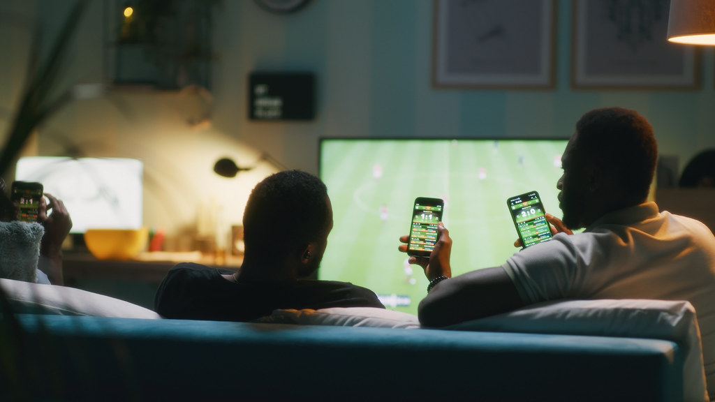 Two men watching football and betting using a mobile phone app.