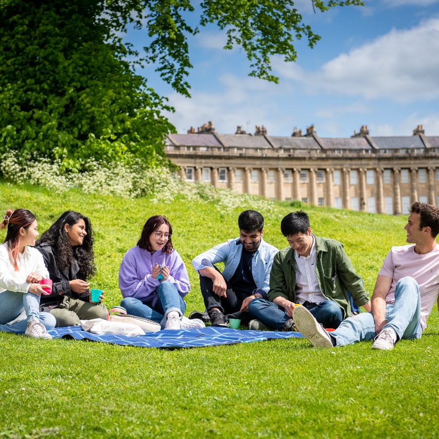 A group of students sit around a blue picnic blanket on the green lawn in front of the Royal Crescent.