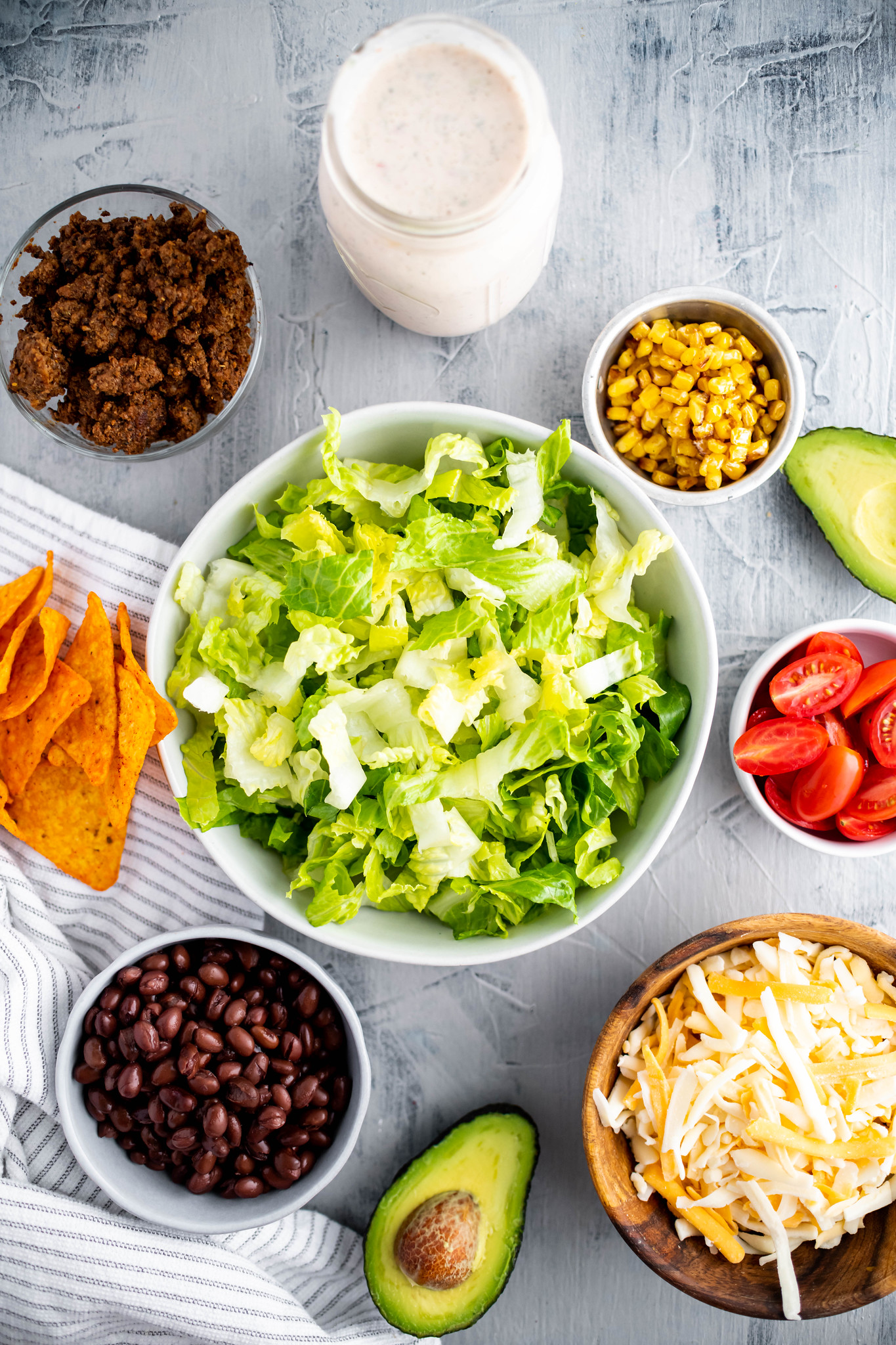Large bowl of chopped romaine lettuce in the center. Surrounded by bowls of corn, shredded cheese, sliced grape tomatoes, corn, black beans and ground beef.