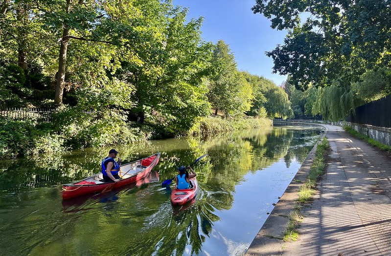 A photo of a canal from the towpath, curving off to the left in the distance. There are trees on either side, lit up by the sun and reflected in the almost still water, along with the clear sky. Two canoeists are in the foreground in red boats heading away from us.