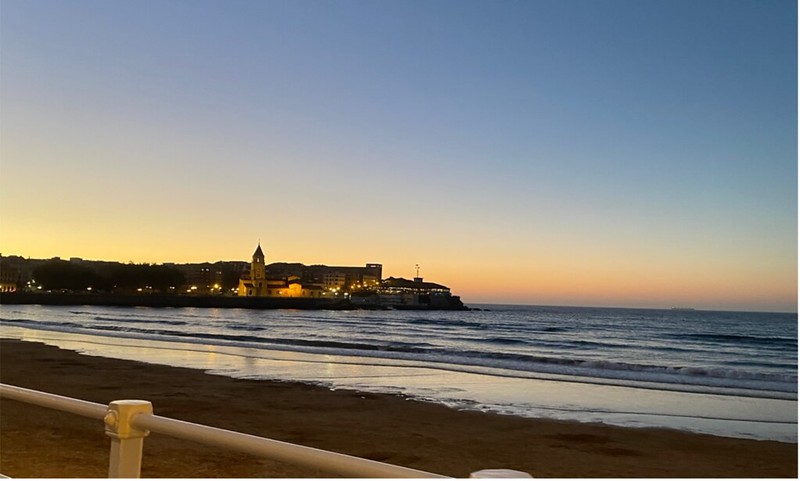 Sky fades from blue to yellow as the sun goes down on the beach with city lights of Gijón in the distance.