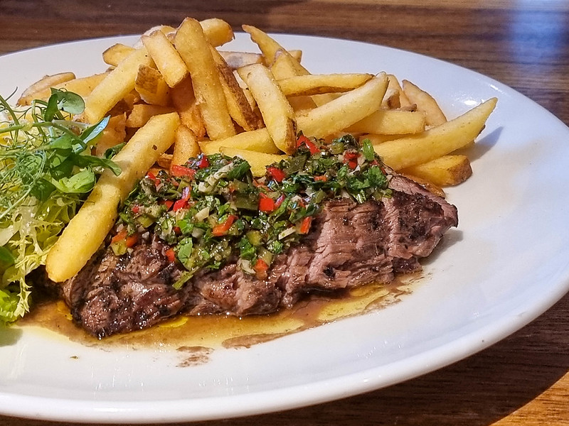 2A steak cut and covered with a green chimichurri sauce. Behind the steak there are French fries.