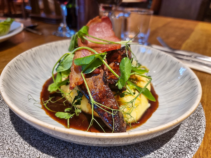 The liver dish: at the bottom of the plate there is the mashed potato, on which there are three pieces of liver. Over there is a slice of bacon and the dish is decorated with small green herbs. At the bottom of the plate there is a brown sauce.