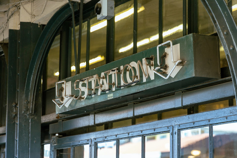 A 'STATION' sign above a door, white on green lettering accented by neon, unlit in the light of day