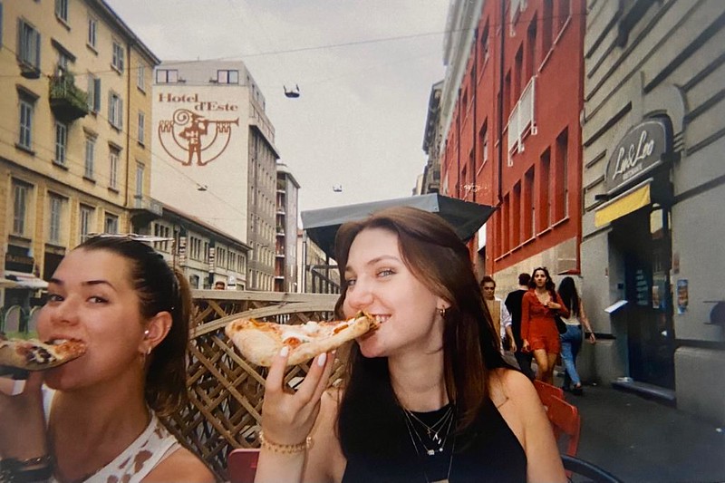 Two girls smiling while biting down on pizza at a restaurant on the street.