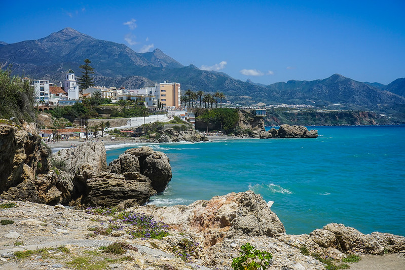 A view over Nerja, with Balcon de Europa in the background. The sea is on the right hand side. There are tall mountains raising behind the town, on the left.