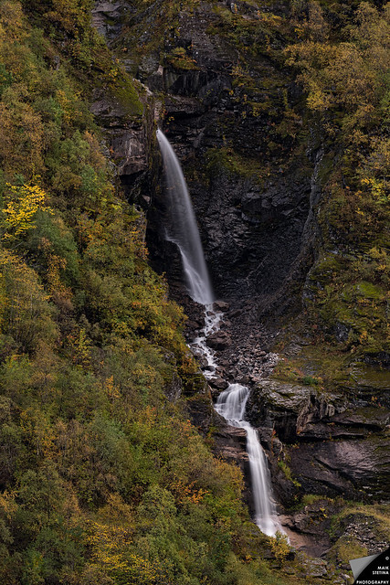 Ein Wasserfall bahnt sich seinen Weg durch den felsigen Berg, vorbei an Bäumen, Sträuchern, Moos und Flechten in herbstlichem Kleid.