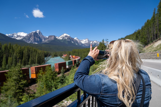 Blonde woman takes photos of a freight train passing through Morant's Curve in Banff National Park on a sunny summer day