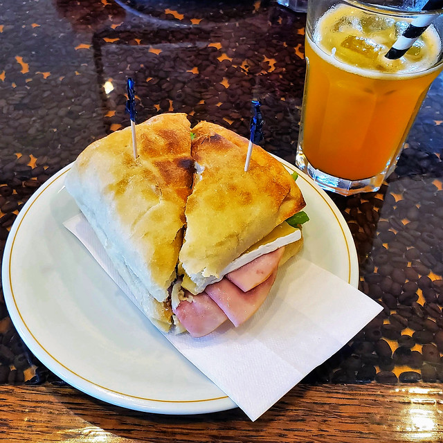 Ham and brie ciabatta bun sandwich; mango / peach soda, on a coffee beans table