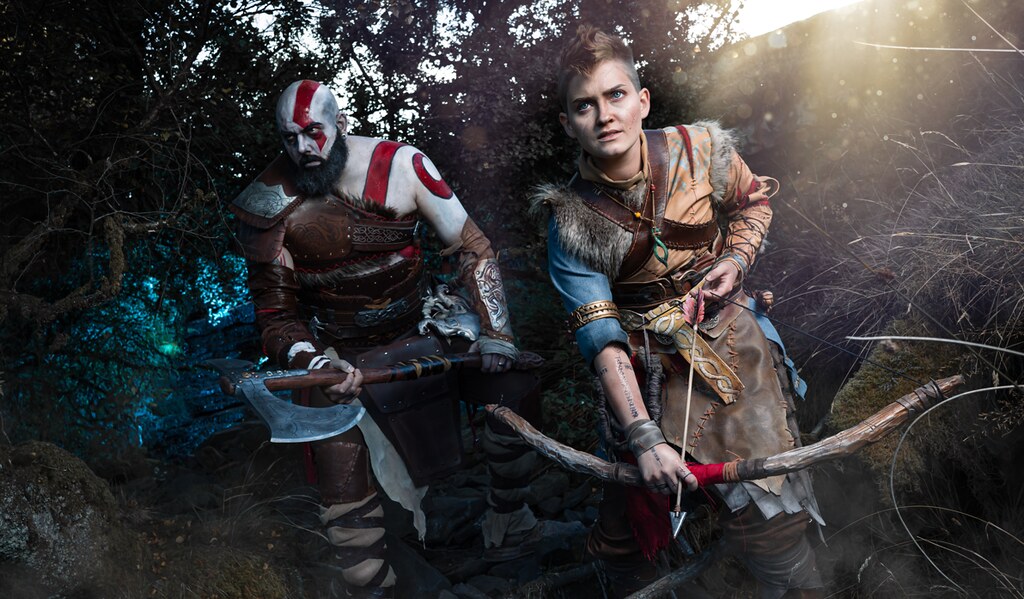 Cette image montre des cosplayers jouant Kratos et Atreus, émergeant d'une forêt. Ils ont l'air parés au combat, leurs armes tirées.