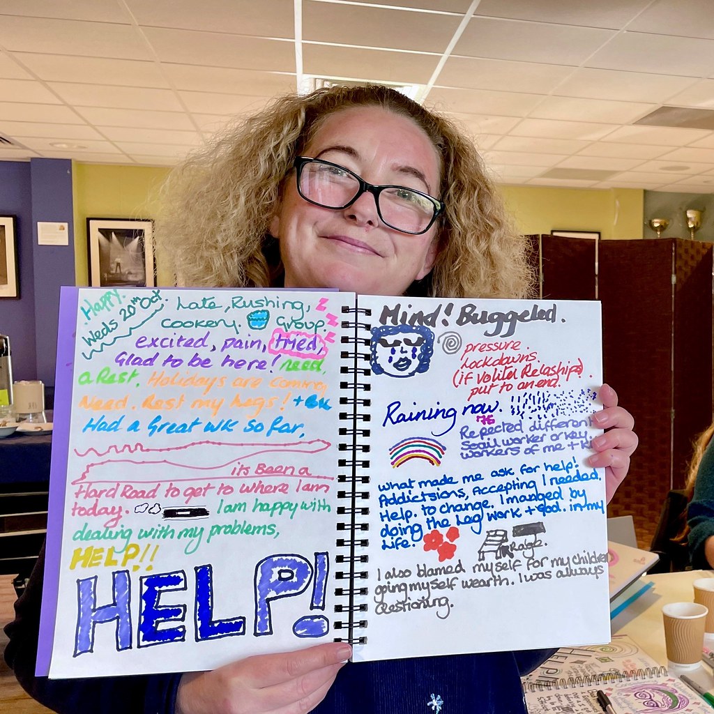 A woman holding an open notebook full of colourful writing and small drawings. The largest word says 'Help'.