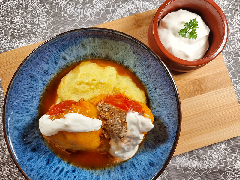 A blue bowl on a wooden board. Inside the bowl there is a yellow pepper cut in half, stuffed with minced meat. It sits on a bed of polenta, and the sauce of the dish is surrounding it. On top of the pepper there is sour cream. Next to it there is a smaller brown bowl with sour cream and a parsley leaf on top.