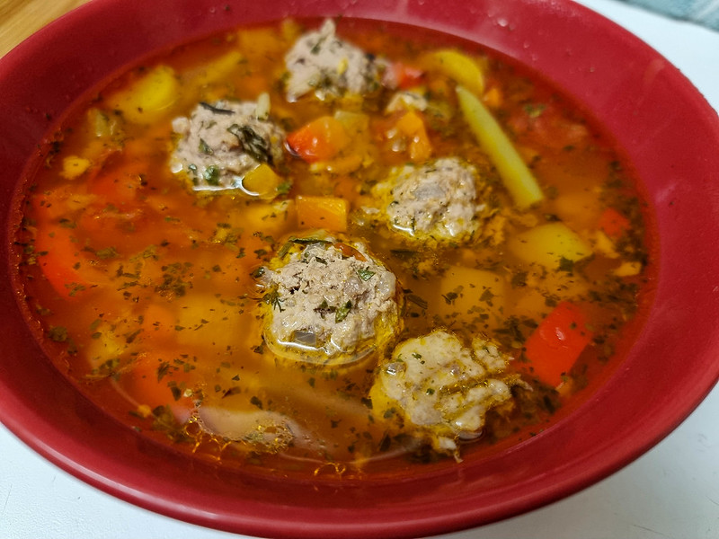 a red bowl filled with the meatballs soup. You can see five meatballs and many vegetables inside the red soup.