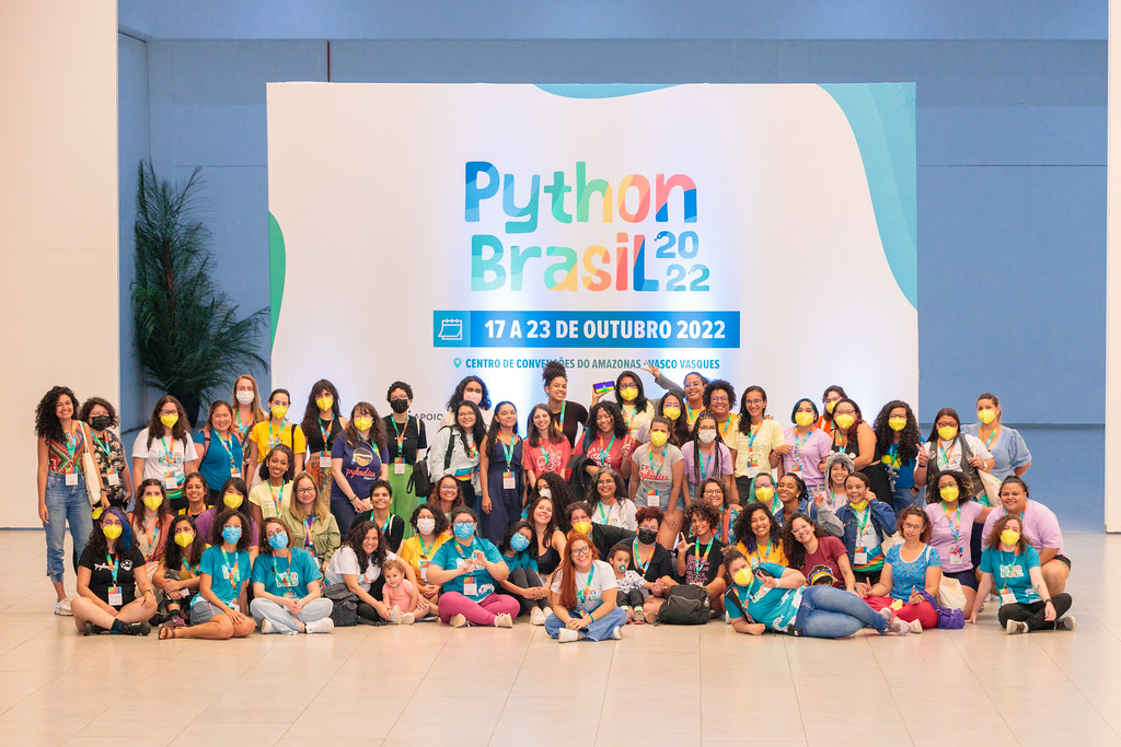 Na imagem estão diversas mulheres da comunidade python em pé e sentadas na frente do banner da Python Brasil 2022