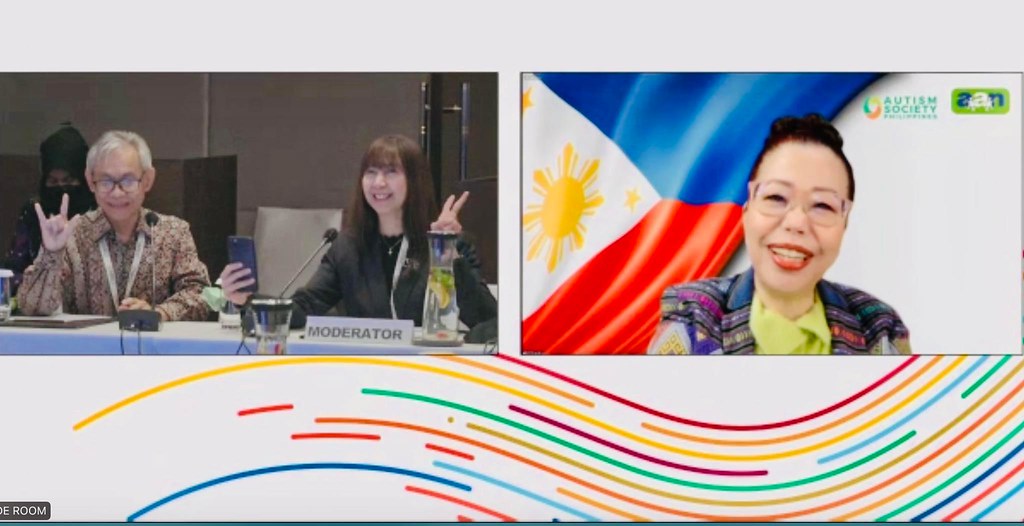 Dang Koe wearing eyeglasses and colorful blouse with a Philippine Flag background. She has black hair. A woman wearing black outfit and she has black hair, doing peace sign and holding her mobile. A man wearing a long sleeved polo and eyeglasses. His hair is color blond.