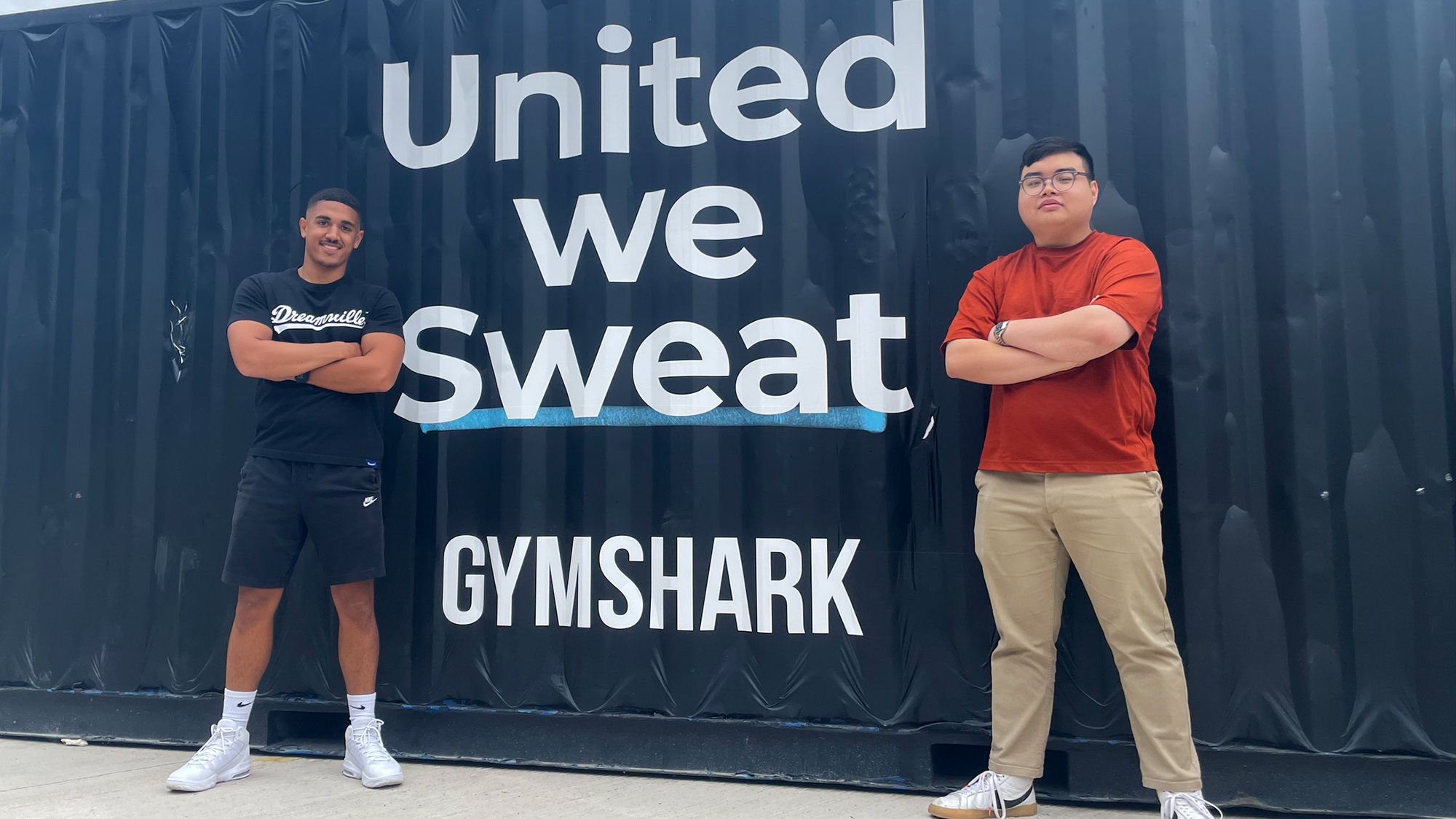 Two young men stood next to the Gymshark logo and strapline 'united we sweat' painted on a dark blue shipping container.