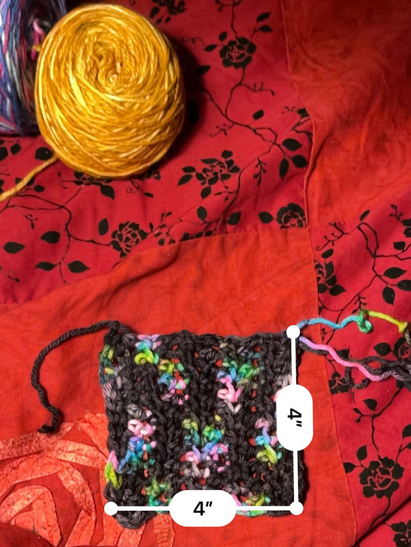 an unwashed, unblocked swatch of worsted weight yarn crocheted in half double crochet; colors are black with spots of bright green/blue/pink; there are 4 inch by 4 inch measurements visible, with dark golden yellow and blue/fuchsia/purple cakes of yarn visible in the top left corner, all on a dark red with roses comforter blanket