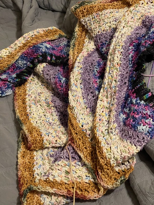 a jumble of crochet fabric in stripes of dark yellow, speckled pink/white/yellow/blue, lilac, variegated blue/fuchsia/purple, and black with specks of bright pink/blue/green, all on top of a messy grey comforter blanket