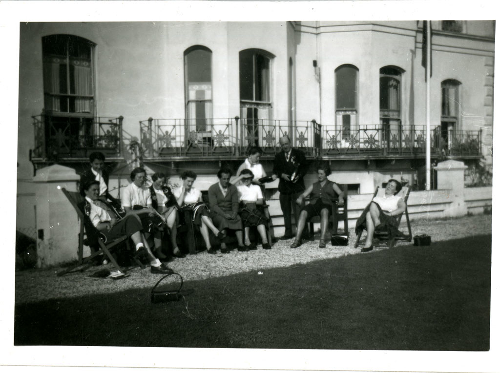 Black and white photo of about 10 ladies and a gentleman sitting in sun in front of building.