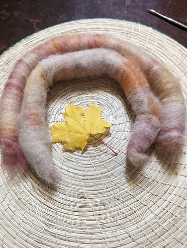 Two handcarded rolags of handdyed alpaca fibre are curled around a yellow maple leaf on a handwoven straw placemat.  The alpaca is prepared by Stone Spindle Farm in Tamworth Ontario.  The placemat is made in Jamaica.