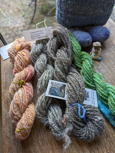 Four skeins of yarn handspun by irieknit lay on a small wooden table.  A mouse knitting a blue sock figurine is to the right of the fourth skein and a handknit handspun toy is behind the mouse.