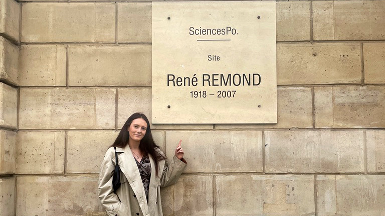 A young woman (Grace) pointing at a sign on the wall outside of The Paris Institute of Political Studies (Sciences Po).