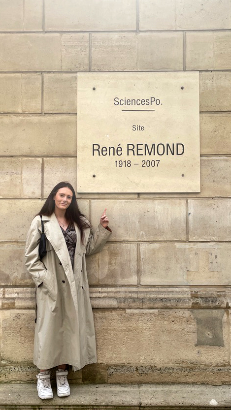 A young woman (Grace) pointing at a sign on the wall outside of The Paris Institute of Political Studies (Sciences Po).