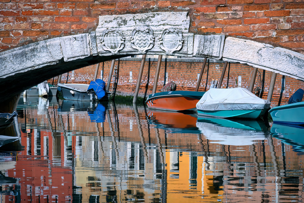 View under a brick bridge of moored boats reflected in the water of a canal, Venice, Italy