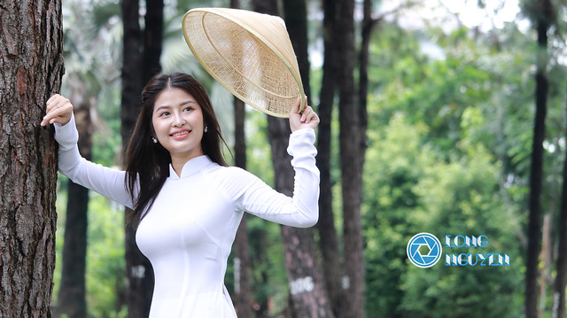 A charming beautiful girl is posing gracefully with Ao Dai white-traditional dress of Vietnam and traditional South East Asian straw rattan conical hat in front of palm trees.