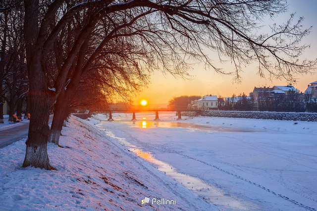 winter sunrise in uzhgorod. beautiful cityscape in early morning. sun above horizon and bridge over the frozen river. snow covered embankment