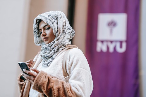 A young woman wearing a head scarf looks at her phone. Behind her is a purple NYU banner - Todo lo que Necesitas Saber Sobre la Beca Pell