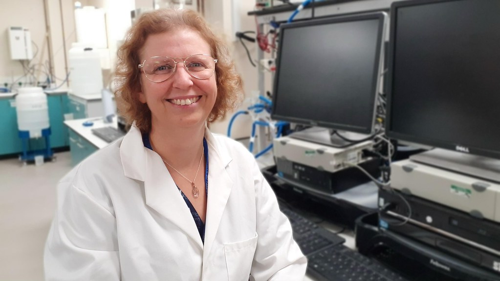 A photo of Dr Christine Edmead, a white woman wearing a lab coat and glasses and smiling at the camera. Behind her is a row of blank monitors.