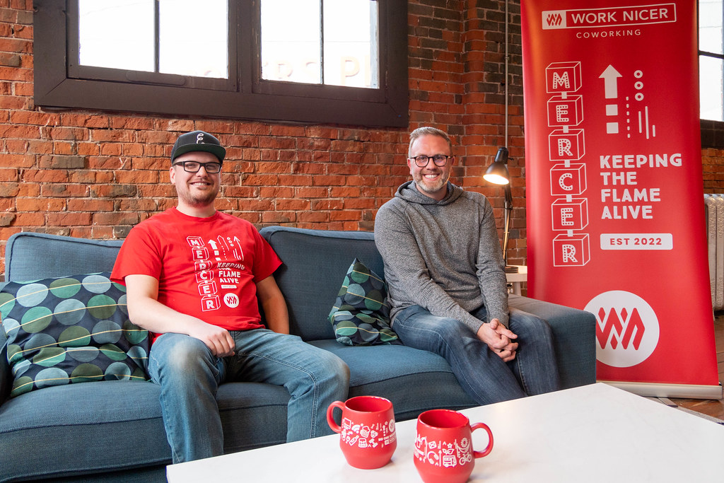 Justin Weleschuk and Alex Putici seated on a couch in front of a brick wall with two red mugs on a white table in front of them
