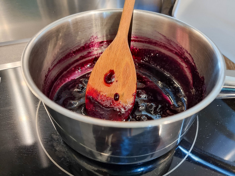 A pot with blueberry jam inside, boiling. There is a wooden spoon inside the pot as well.