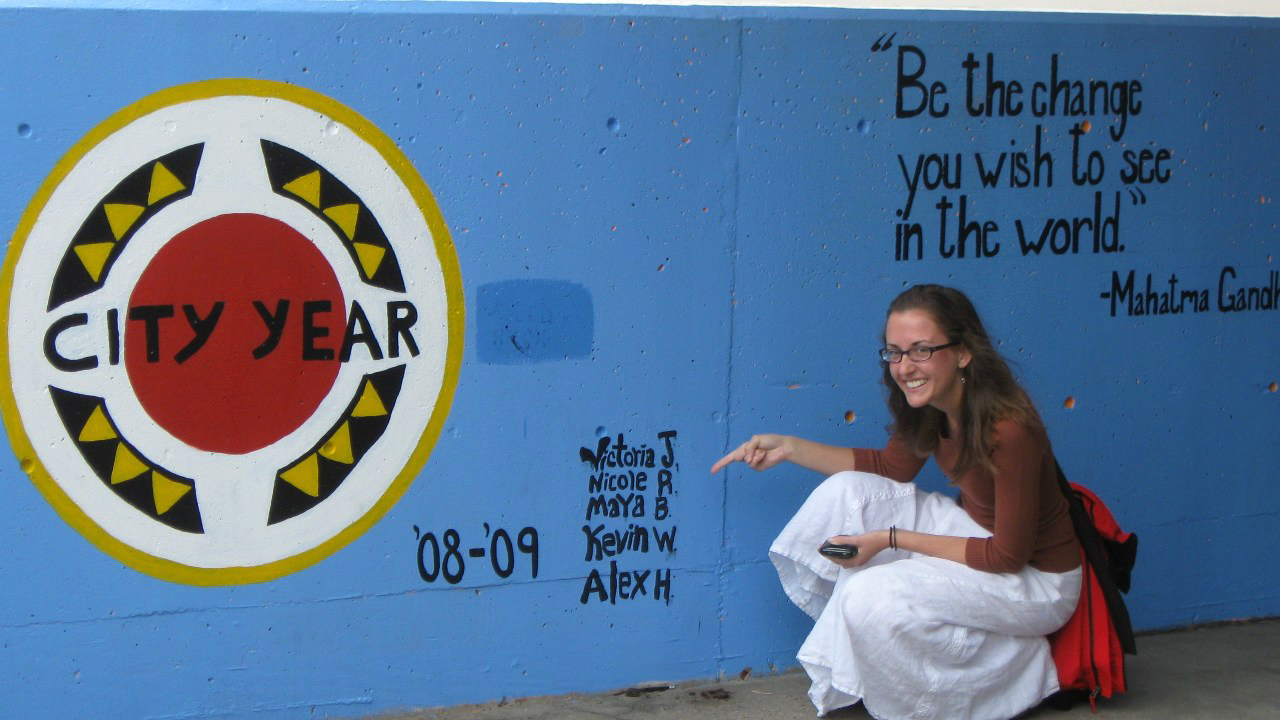 A young woman (Maya Butovskaya) smiling in front of a painted mural that says 'City Year' on it.