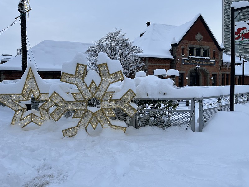 Large decorative snowflake and star with snow piled up on them in front of a restaurant patio with snow piled up on the tables and heaters, which is in front of a restaurant with snow on the roof. Photo taken in New Westminster on Dec 20, 2022