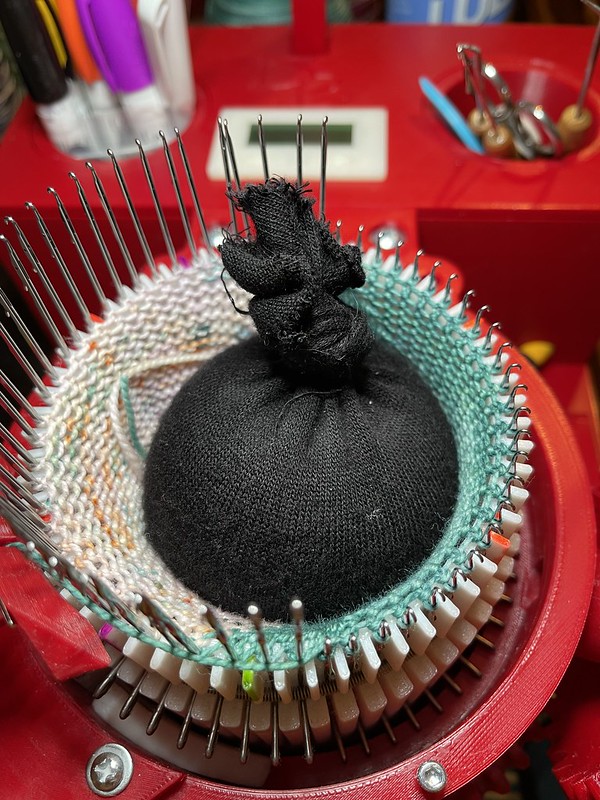 a circular sock knitting machine with needles raised for working the heel, and a soft round weight in the center; the yarn is teal for the heel and speckled off white, orange, and teal for the main part of the sock