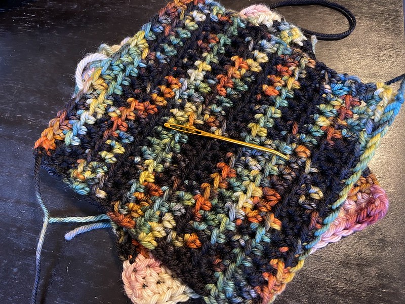 stack of crochet pockets with a needle on top; yarn on the top square is black with burnt orange, faded teal, and dark yellow; underneath, some cranberry and cream colors are visible
