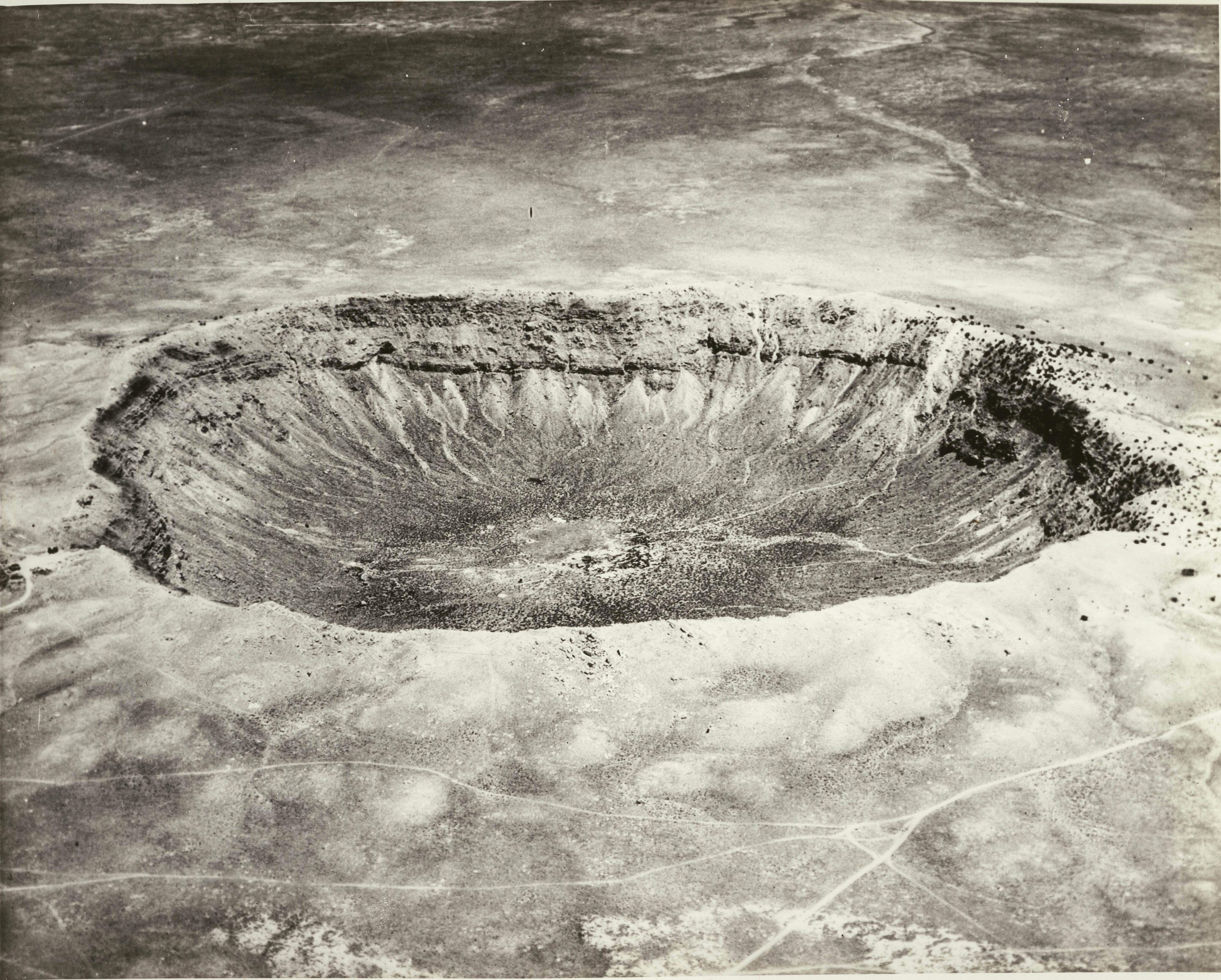 Photograph of the Barringer Crater in Arizona, ca. 1920. A large depression is pictured at center, striated with different colors of sand and other mineral deposits. The surrounding area appears to be barren and flat. | src USC