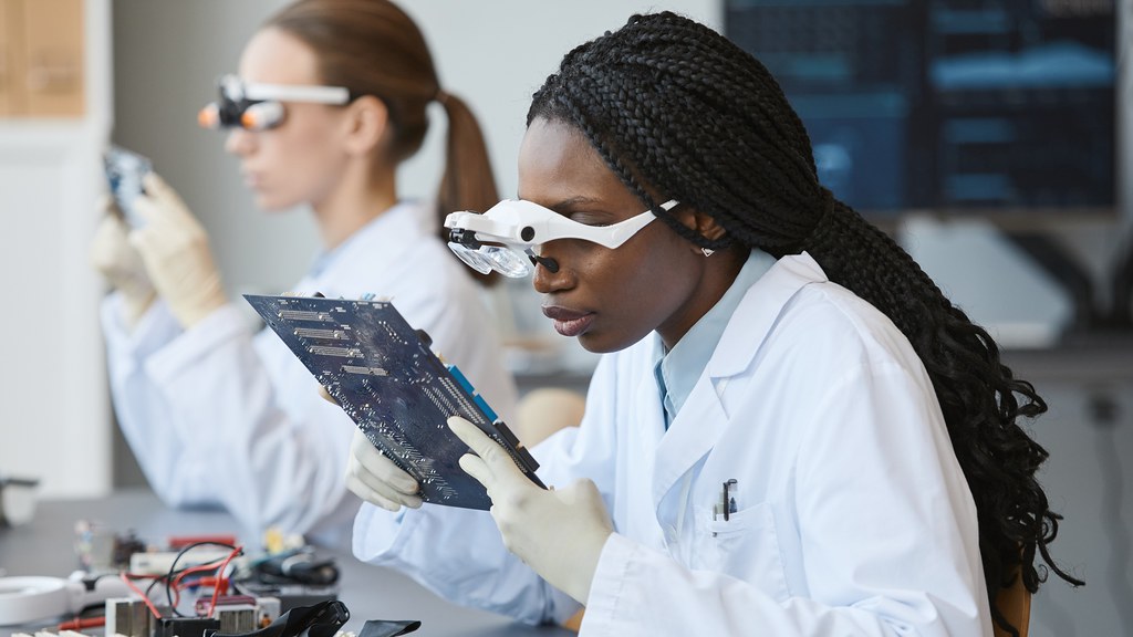 A young woman in a white coat examines electrical components