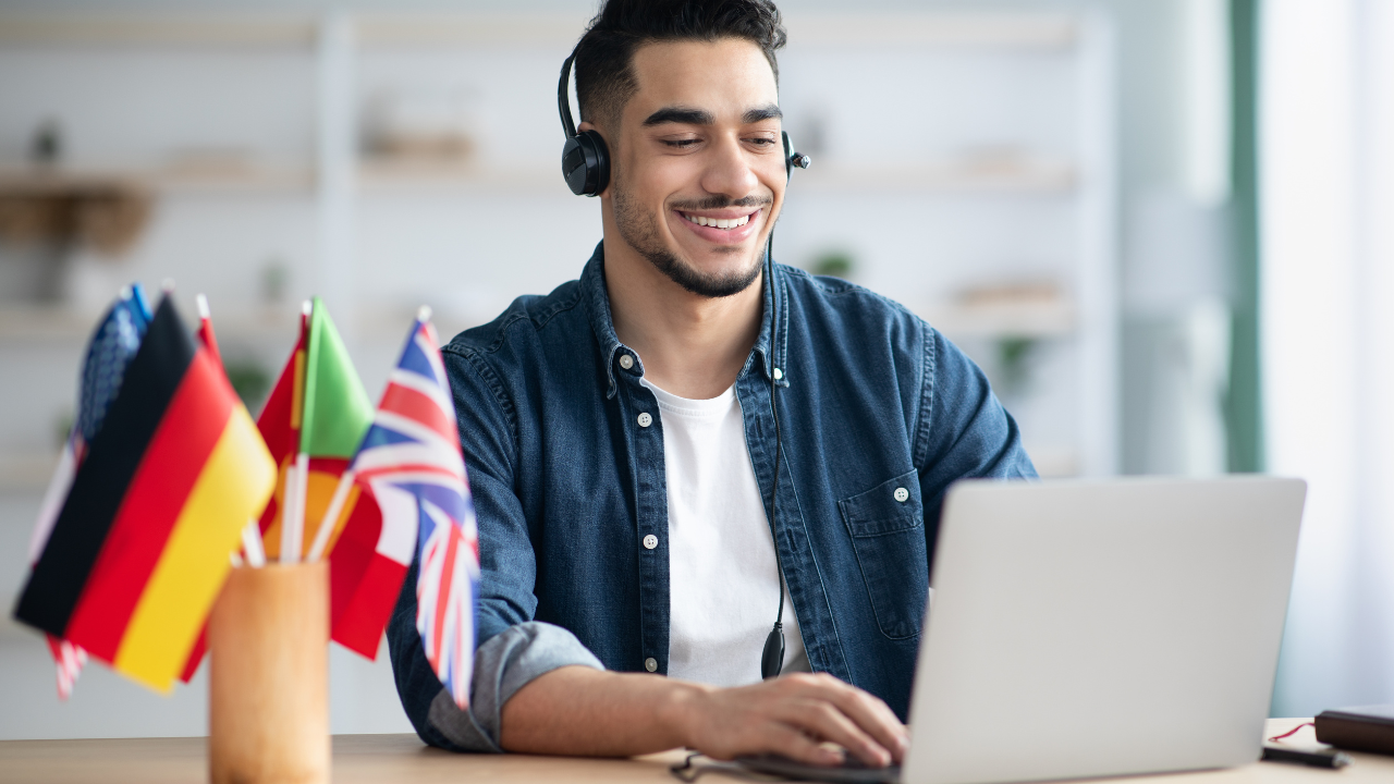 Smiling student wearing headphones learning a foreign language on a laptop. On the desk are various country flags.