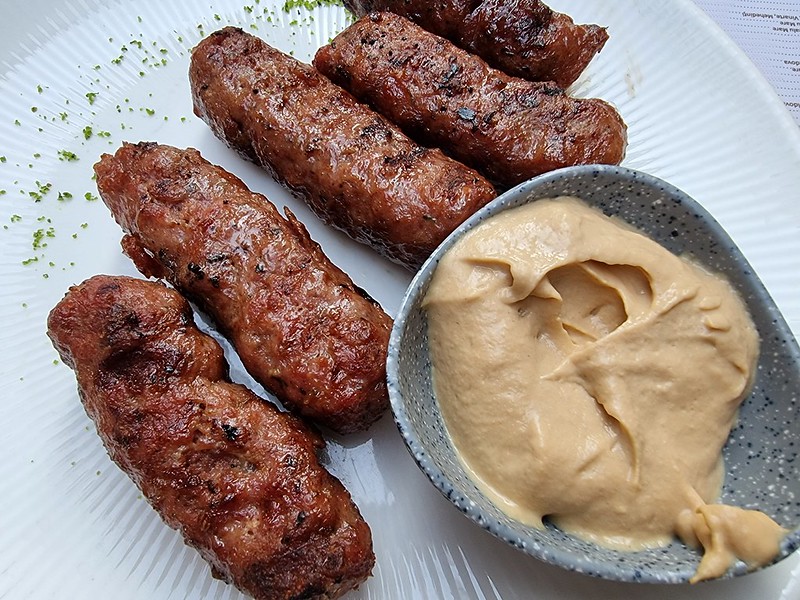Five skinless grilled sausages next to a grey ramekin filled with yellow mustard.