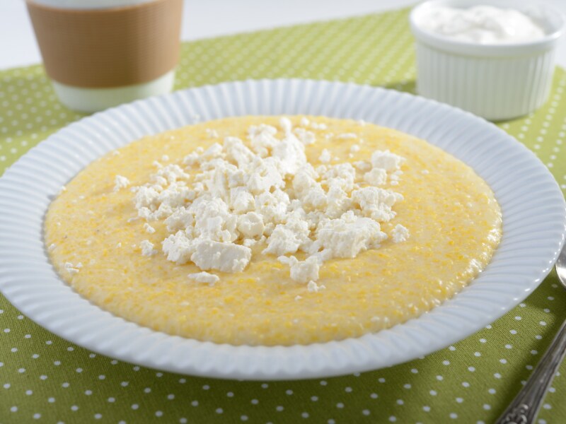 a white plate filled with soft yellow polenta. There is grated white soft cheese on top. Behind there is a white ramekin filled with sour cream.