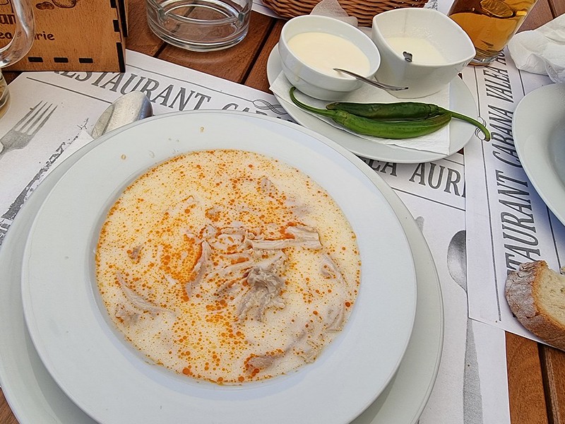 A white deep soup filled with a yellow soup. You can see the slices of tripe inside the soup. Behind the plate, on the same table, there is a plate with two white bowls, one filled with garlic sauce and the other one with sour cream. Next to them there are two green chili peppers