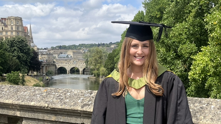 A young woman wearing a ceremonial gown and mortarboard while stood on a bridge in front of buildings in Bath city centre.