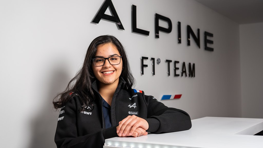 Female smiles to camera in front of wall with Alpine F1 logo on it